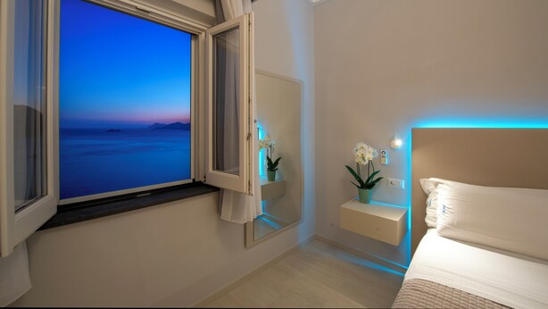 A bedroom with a large window, a bedside table with a plant, and a bed with white bedding. - House Salú Suite (Praiano)