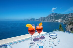 Two glasses of red beverage with orange slices on a table with a decorative border, overlooking a coastal landscape with mountains and the sea. - House Salú Suite (Praiano)