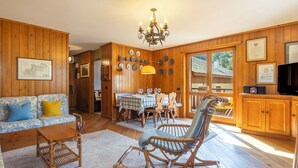 Ein Holzinterieur mit Esstisch, Stühlen, einer Couch, einem Schaukelstuhl, einem Fernseher und an Wänden befestigten Tellern.
