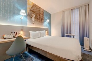 Habitació d'hotel amb llit, escriptori, cadira, làmpada i una finestra gran amb cortines.