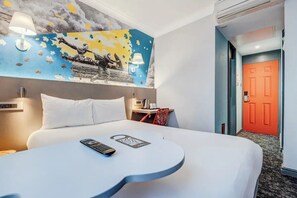 Et hotelværelse med en seng, et lille bord med en fjernbetjening, en lampe på væggen og en rød dør i baggrunden.