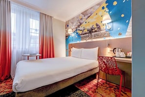 Habitación de hotel con una cama, una silla roja, una mesita y una pared decorada con un mural, además de una ventana con cortinas.