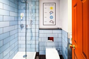 Et badeværelse med blå fliser, en glasbrusekabine, en håndsæbeautomater og en plakat med vejrsymboler.