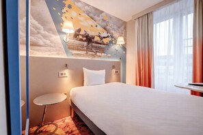 Et hotelværelse med en seng, et lille natbord, en lampe på væggen og et vindue med gardiner.