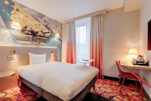 Habitación de hotel con cama, escritorio, silla, mesita, lámpara y un mural en la pared.