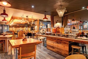 Un restaurante con mesas y sillas de madera, una zona de bar con taburetes, iluminación colgante y un candelabro.