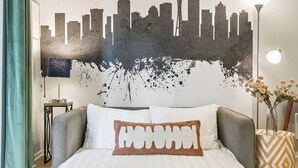Un dormitorio con cama, un mural de paisaje urbano, una lámpara y un florero con flores.