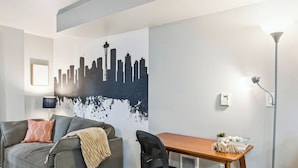 Una sala de estar moderna con un sofá gris, una mesa de madera, una silla de oficina negra, un mural con un horizonte urbano y una lámpara de pie.