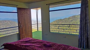 A bedroom with a bed, a wooden door, and large windows offering a view of a hilly landscape. - Kanatal Greenhood Resort (Kanatal)