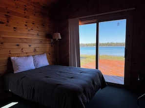 A wooden cabin room with a bed, a lamp, and a large window overlooking a lake. - Drummond Island Hotel & Vacation Homes - Kaye's Little Cabin (Drummond)