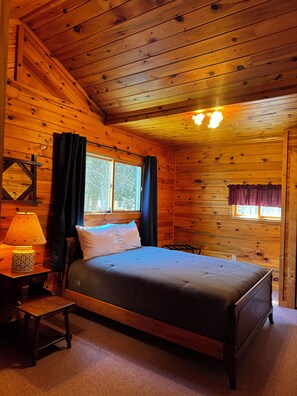 A wooden cabin bedroom with a bed, a nightstand, a lamp, and a window with curtains.