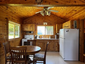 A wooden cabin kitchen with a stove, refrigerator, wooden cabinets, a dining table with chairs, and a ceiling fan.