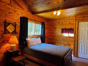 A wooden cabin room with a bed, a nightstand, a lamp, a window with curtains, and a door.