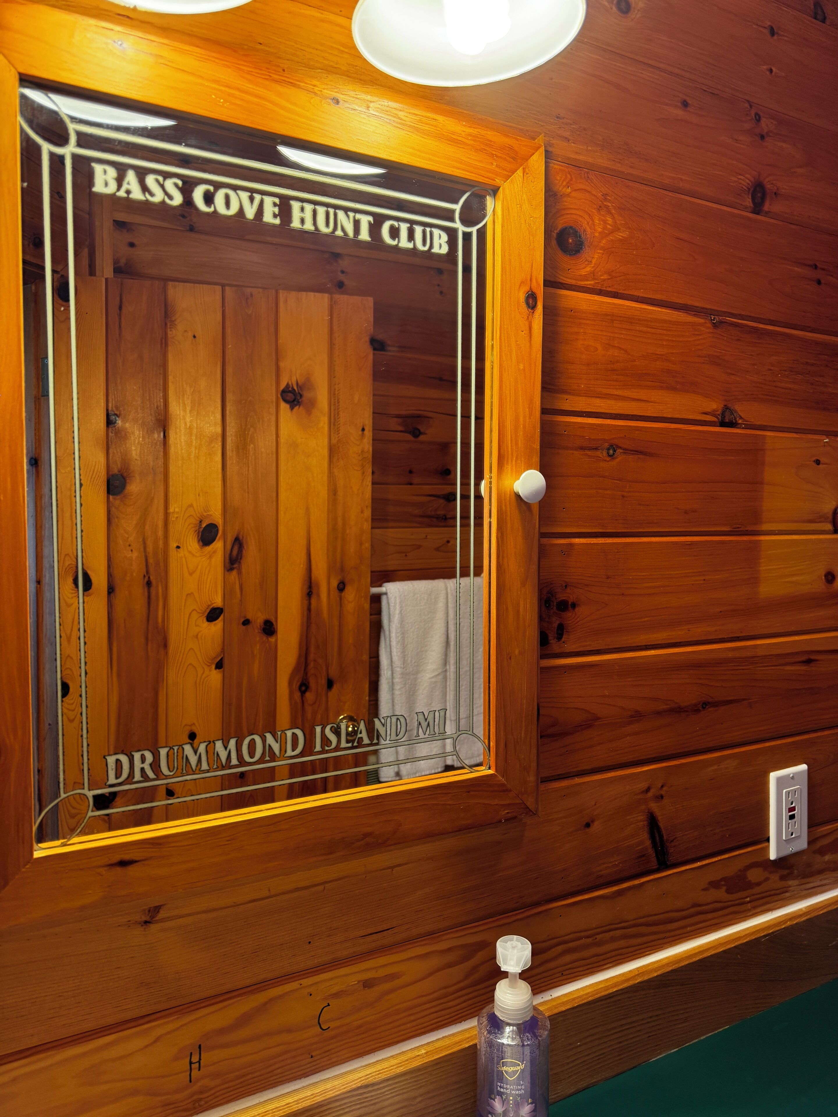 Een houten deur met een spiegel, waarop 'Bass Cove Hunt Club' en 'Drummond Island MI' staat.