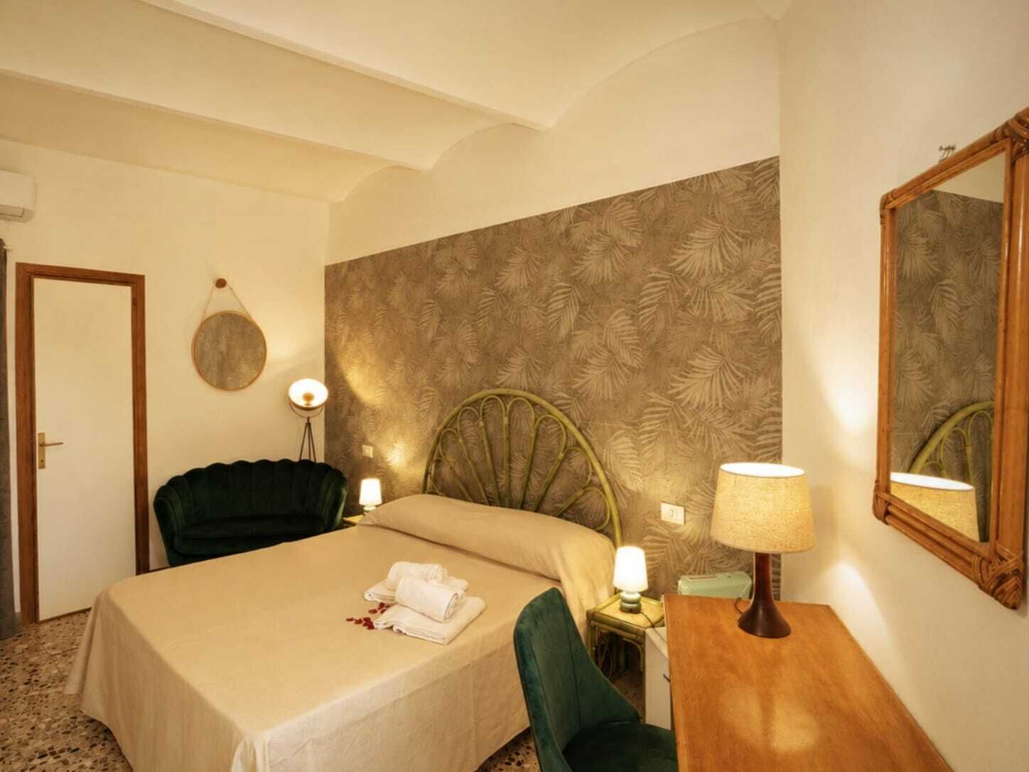 Une chambre d’hôtel comprenant un lit, une chaise, un bureau, une lampe, un miroir et un mur orné d’un motif de feuilles.