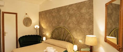 Quarto de hotel com cama, cadeira, escrivaninha, luminária, espelho e parede com padrão de folhas.