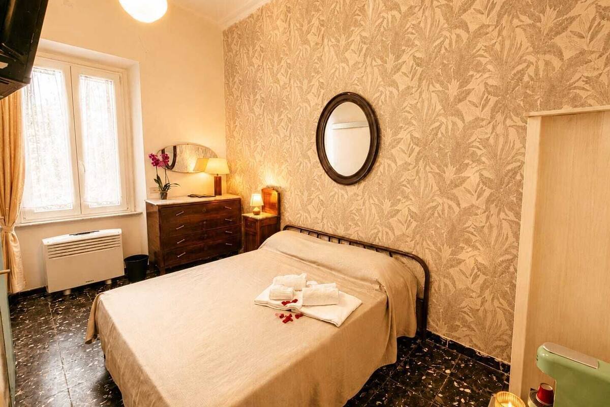 Una habitación de hotel con cama, cómoda, espejo, ventana con cortinas, radiador y pared con estampado floral.