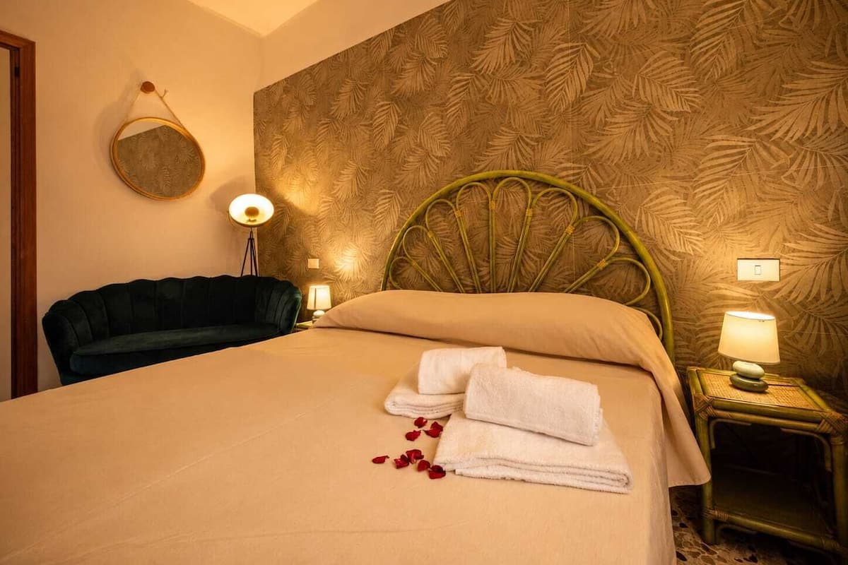 Habitación de hotel con una cama, un espejo redondo, cabecera verde, dos lámparas de noche, una silla verde y una mesita con lámpara.