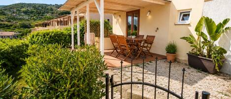 A covered outdoor seating area with a table and chairs, surrounded by greenery and a gravel path leading to a gate.