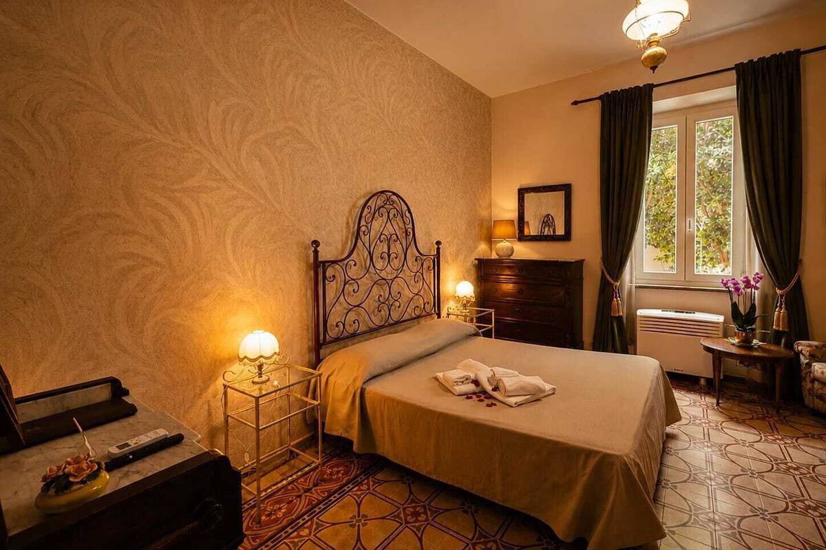 Habitación de hotel con cama, lámparas de noche, cómoda, ventana con cortinas y una mesita con un jarrón de flores.