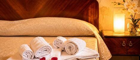 A neatly made bed with rolled towels and rose petals, a bedside table with a vase of flowers, and a lamp.