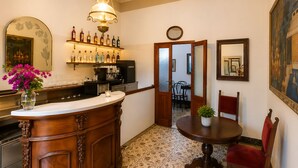 A bar area with a wooden counter, a coffee machine, and a variety of bottles. There is a dining area with a round table and chairs, and a large painting on the wall. - Boutique Hotel Golfo Azzurro (Capoliveri)