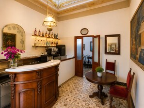 Una zona de bar con mostrador de madera, una máquina de café y varias botellas. Hay un comedor con una mesa redonda y sillas, y un cuadro grande en la pared.