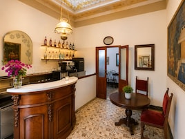 A bar area with a wooden counter, a coffee machine, and a variety of bottles. There is a dining area with a round table and chairs, and a large painting on the wall.