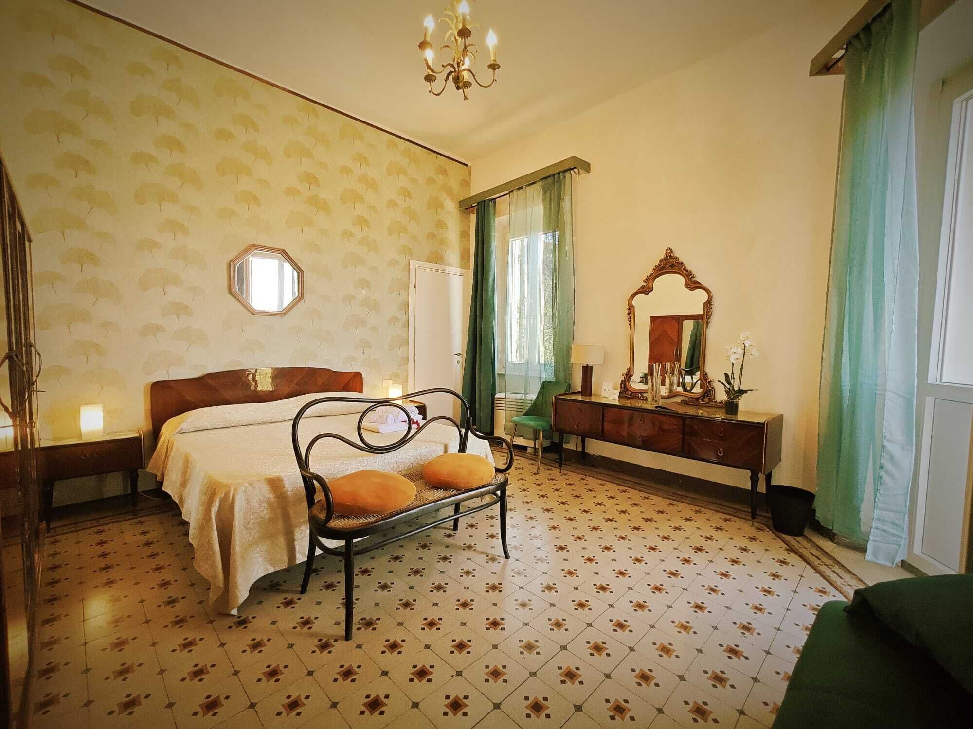 A hotel room with a bed, a bench with cushions, a dresser, a mirror, and a chandelier.
