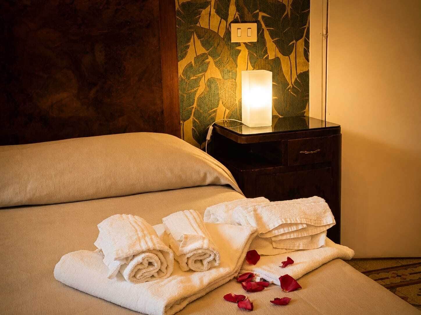 Una cama bien hecha con toallas blancas enrolladas y pétalos de rosa, una mesita de noche con lámpara y una pared con diseño de hojas.