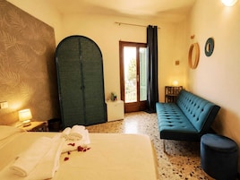 A hotel room with a bed, a blue sofa, a large arched door, a small table with a lamp, and a window with curtains.