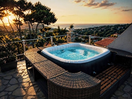 A hot tub on a stone deck with wicker furniture, overlooking a scenic landscape.
