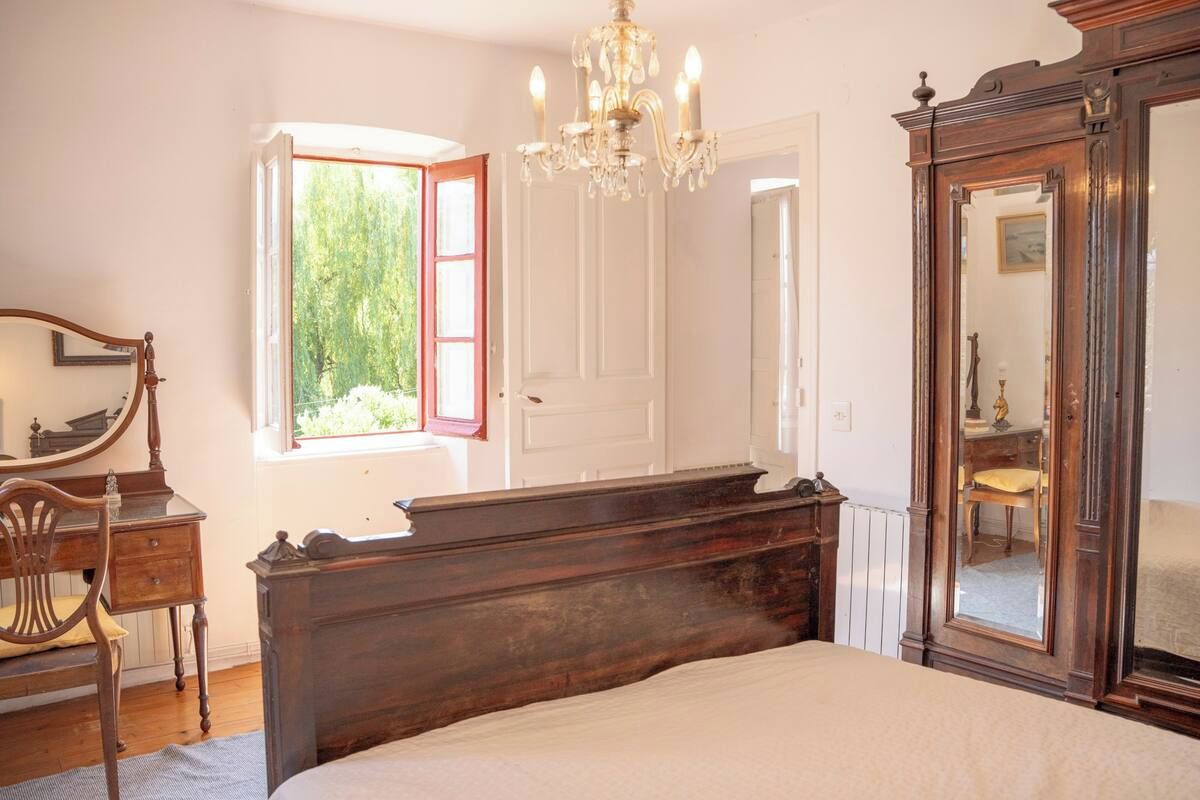 Ein Schlafzimmer mit einem großen Bett, einer Holzkommode, einem Spiegel und einem Fenster mit Blick auf Grünflächen.