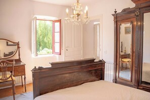 A bedroom with a large bed, a wooden dresser, a mirror, and a window with a view of greenery.