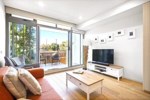 Ein modernes Wohnzimmer mit Sofa, Fernseher, Couchtisch und einer Schiebeglastür, die zu einem Außenbereich führt.