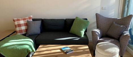 Una sala de estar con sofá, sillón, reposapiés y una mesita de centro con un libro.