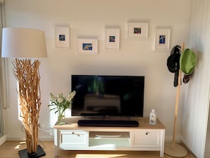 Ein Wohnzimmer mit Fernseher, gerahmten Bildern, einer Lampe, einer Pflanze und einem Schuhregal.