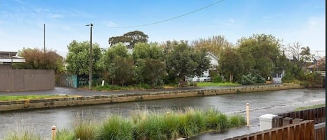 A river with a wooden walkway, trees, and a residential area in the background.
