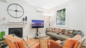 Een woonkamer met een open haard, een flatscreen televisie, een bank met bloemenkussens en een groot raam met uitzicht op bomen.