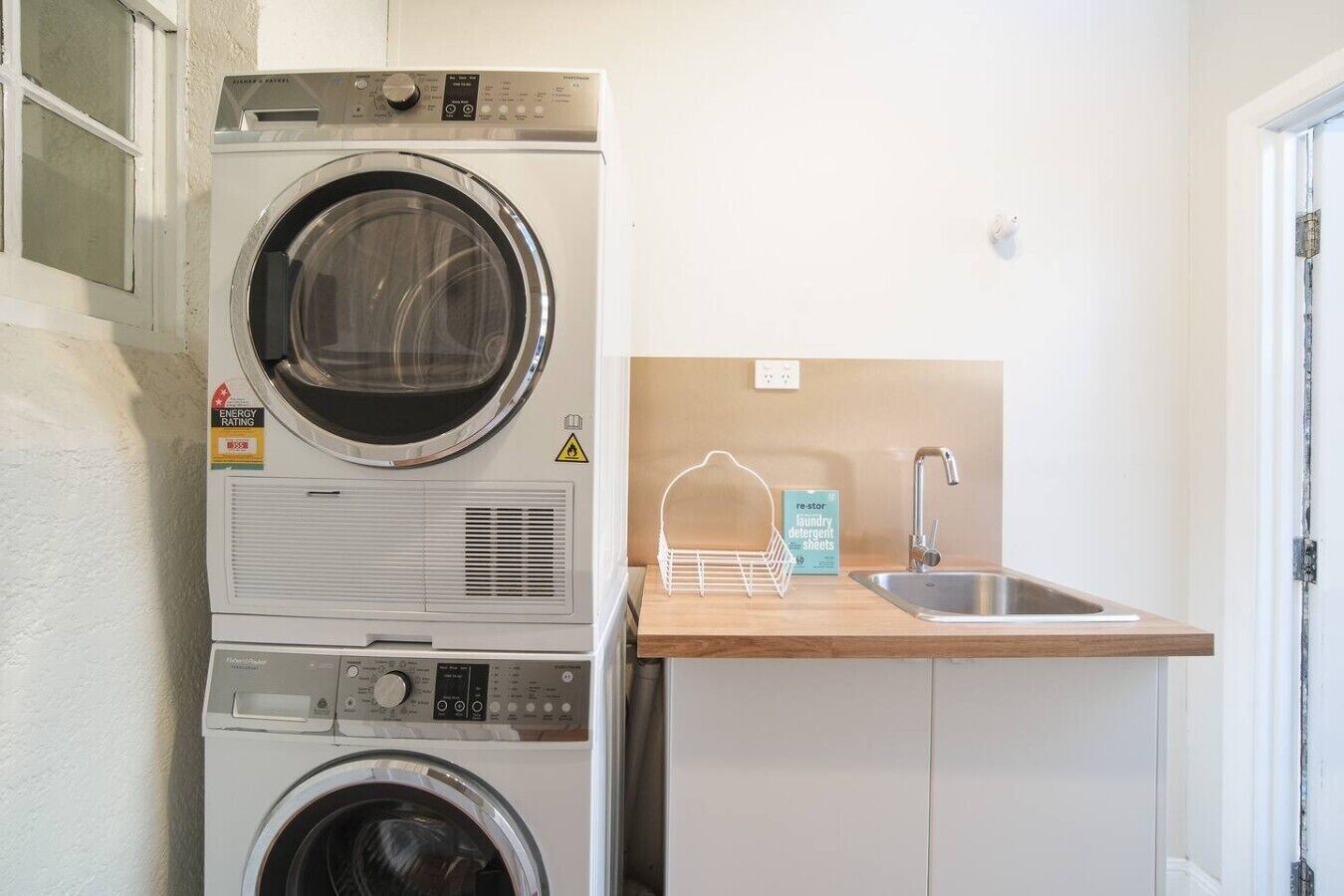 Un espace buanderie équipé d’une pile de lave-linge et de sèche-linge, d’un évier et d’une étagère où sont rangées les lessives de la marque « Re-Store ».