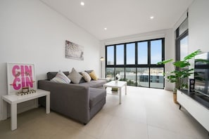 A modern living room with a grey sofa, a small white table, and a large window offering a city view. - Cozy 2B2B near CBD & Airport-w/parking & pets (Rosebery)
