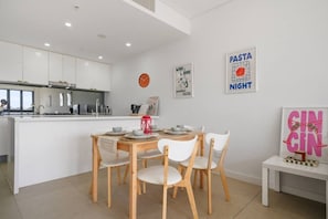 A modern kitchen with white cabinets, a dining area with a wooden table and white chairs, and wall art including 'Pasta Night' and 'Wine & Dine' posters. - Cozy 2B2B near CBD & Airport-w/parking & pets (Rosebery)