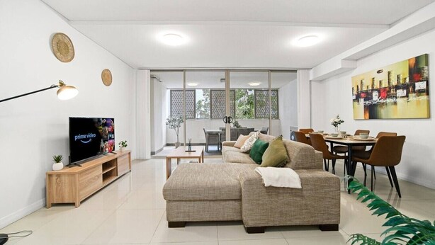 A modern living room with a sectional sofa, a TV, a dining area with a table and chairs, and a large window. - Spacious Mascot 2BR Near Airport + Parking/Pets (Mascot)