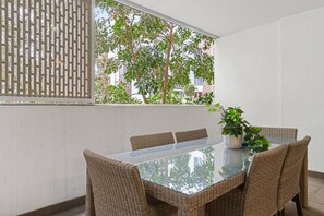 Ein Essbereich mit einem Tisch mit Glasplatte, Korbstühlen, einer Topfpflanze und Blick auf Bäume und Gebäude durch ein Fenster.