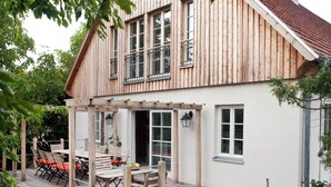 A two-story wooden house with a covered patio, outdoor seating, and a garden.