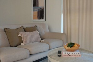 A living room with a sofa, a round coffee table, a remote control, a book titled 'IDEA ALPH,' and a framed abstract painting on the wall. - Cosy Family Retreat | Minutes to Hospital & Woden (Garran)