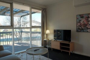Ein Zimmer mit Fernseher auf einem Holzständer, einer Lampe, einem runden Tisch und einem großen Fenster mit Blick auf eine Terrasse und Bäume.
