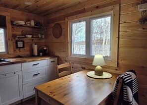 Interior de una cabaña de madera con cocina, mesa de comedor, sillas, lámpara y ventana con vistas a árboles.