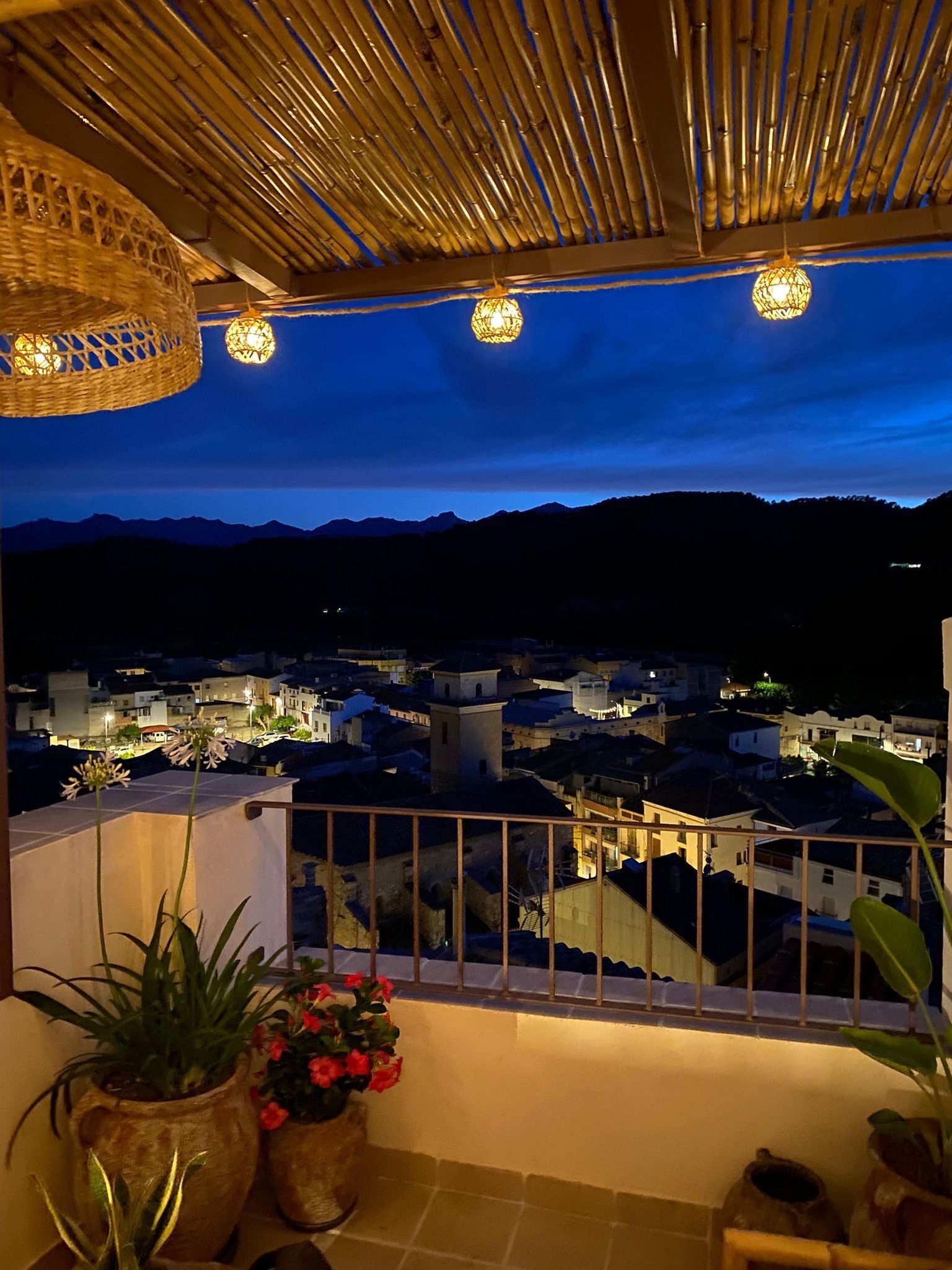 A balcony with a view of a cityscape at dusk, potted plants, and a bamboo ceiling.