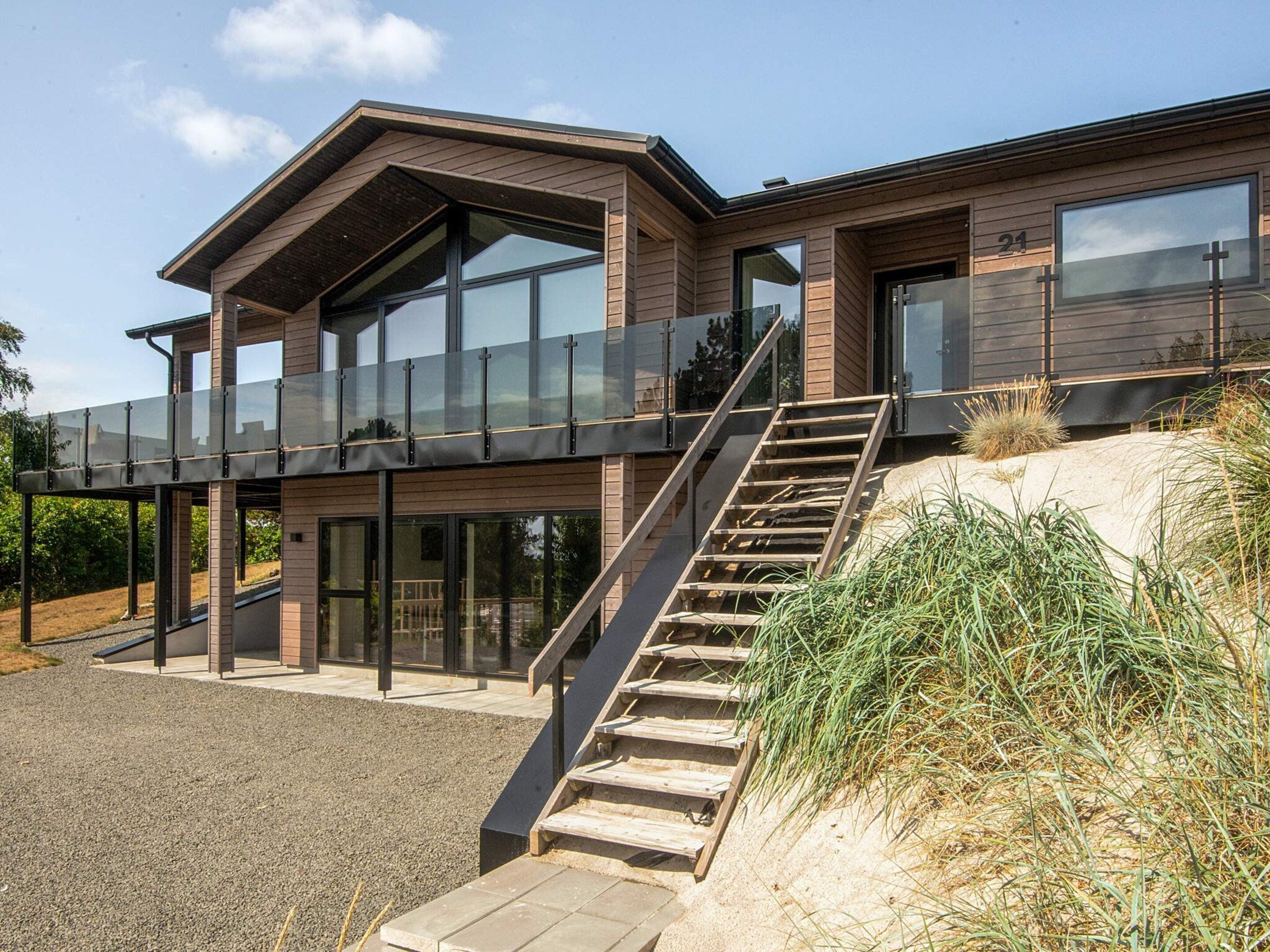 A modern two-story house with a glass balcony, a wooden staircase, and a sandy area with grass.